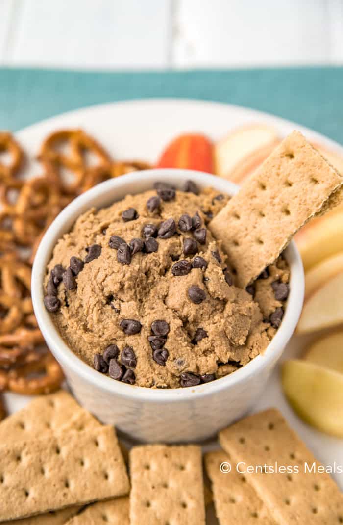 Edible cookie dough in a white bowl with a cracker being dipped in