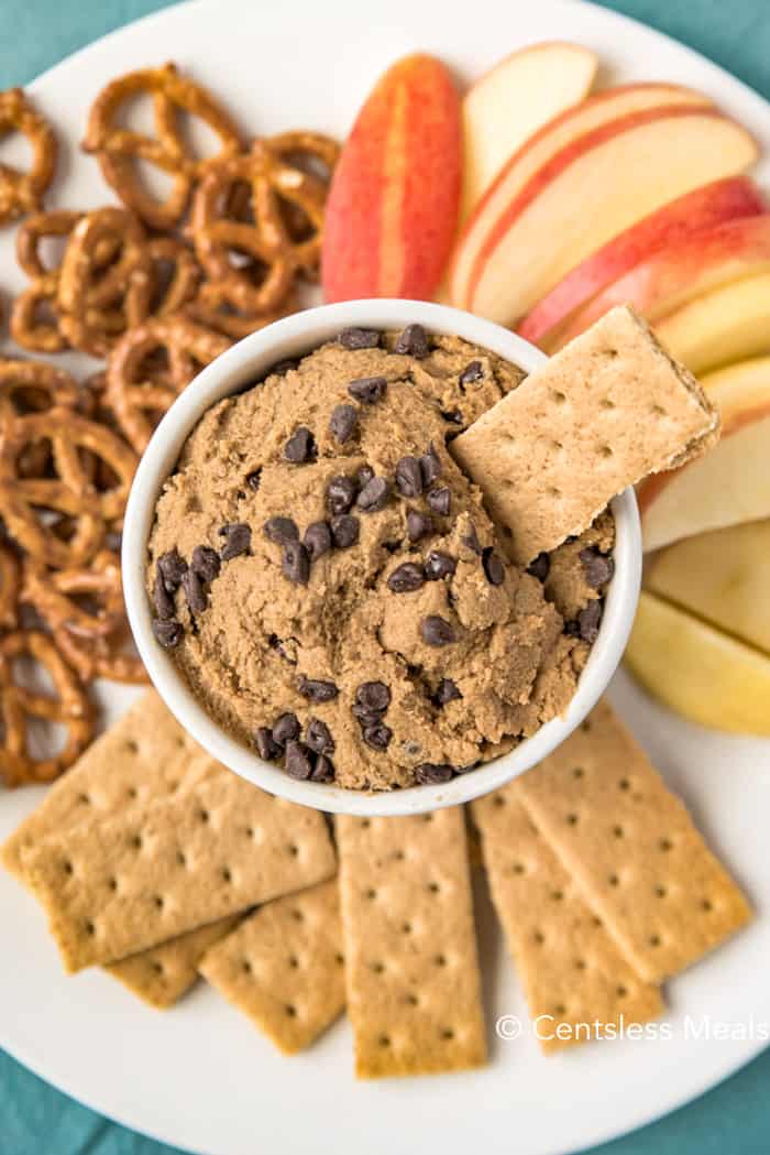 Edible cookie dough in a white bowl with crackers pretzels and apples