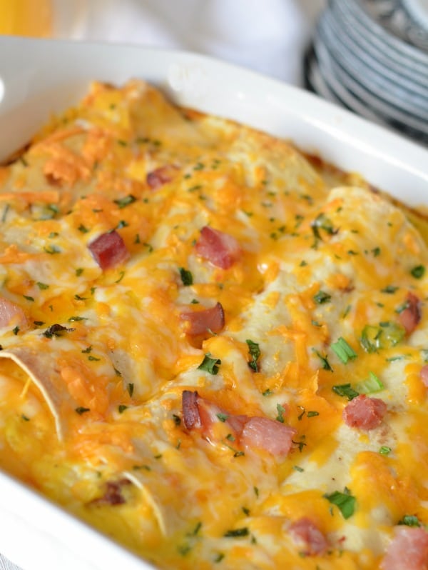 Overnight breakfast enchiladas in a white casserole dish with green onions parsley and ham as garnish
