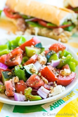 Easy Greek salad on a white plate with a Subway sandwich in the background