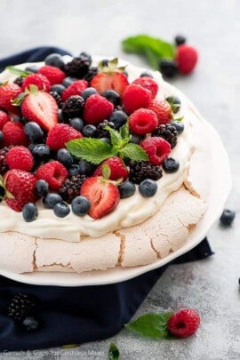 Pavlova topped with berries on a white plate with berries garnished with mint leaves.