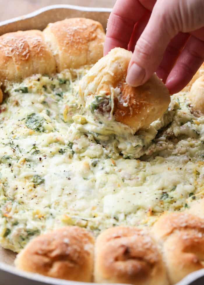 Pull-A-Part crescents with spinach artichoke dip in a skillet with a piece of bread being dipped into the spinach dip