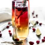 Cranberry Champagne cocktail being served on a round silver tray garnished with fresh cranberries, lime wedges, and rosemary sprigs.