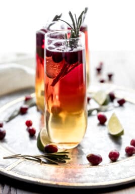 Cranberry Champagne cocktail being served on a round silver tray garnished with fresh cranberries, lime wedges, and rosemary sprigs.