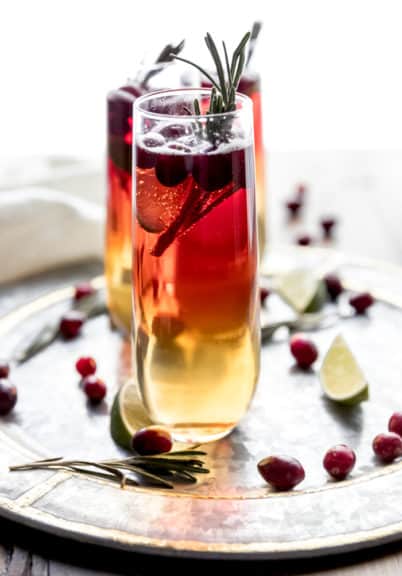 Cranberry Champagne cocktail being served on a round silver tray garnished with fresh cranberries, lime wedges, and rosemary sprigs.