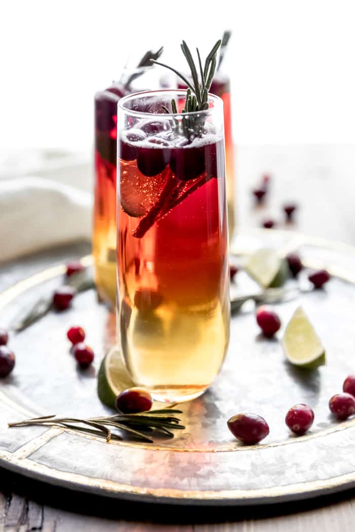 Cranberry Champagne cocktail being served on a round silver tray garnished with fresh cranberries, lime wedges, and rosemary sprigs.