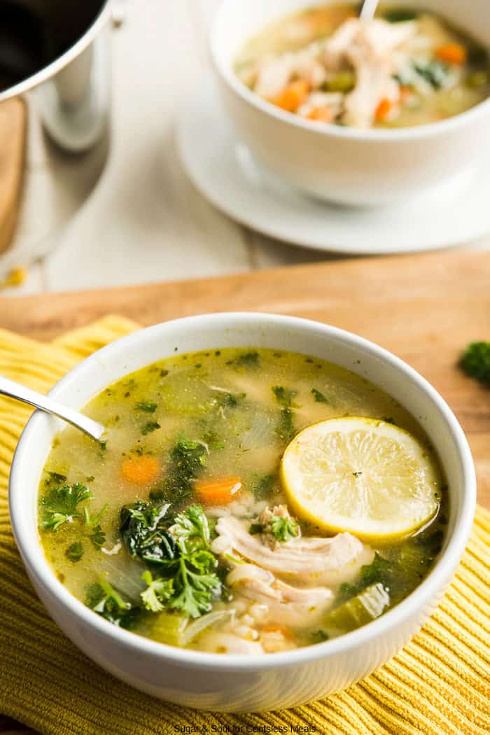 Chicken kale soup in a white bowl with a spoon and a lemon slice