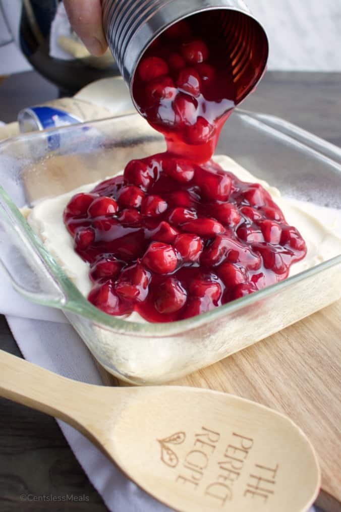 Canned cherries being poured into a casserole dish for cheesecake bake