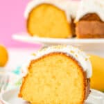 Slice of orange bundt cake on a white plate with a pink background and more cake on a cake stand.