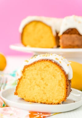 Slice of orange bundt cake on a white plate with a pink background and more cake on a cake stand.