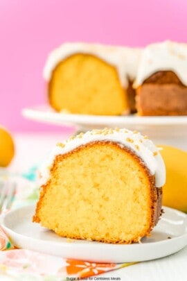 Slice of orange bundt cake on a white plate with a pink background and more cake on a cake stand.