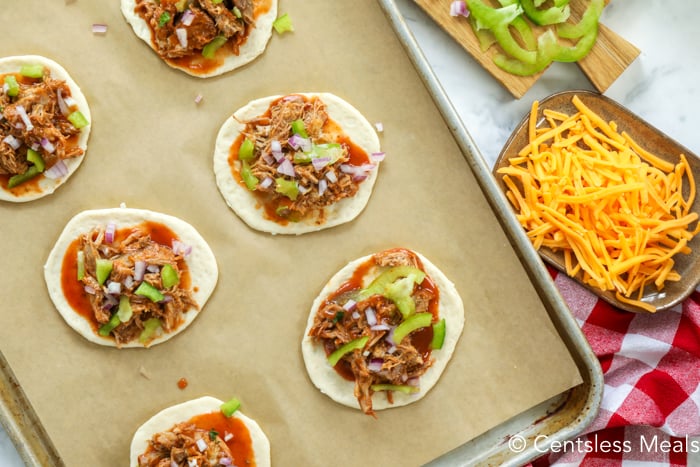 Raw pulled pork mini pizzas on a sheet pan with cheese on the side