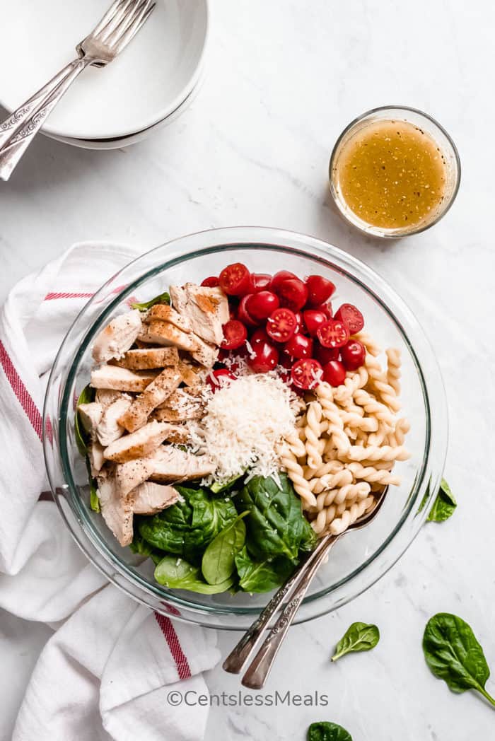 Ingredients for Spinach and Tomato Chicken Pasta Salad grouped together in a clear bowl, with the dressing mixed on the side.