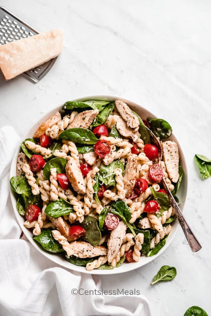 Spinach and Tomato Chicken Pasta Salad served in a white bowl with a silver spoon, sitting on a white background