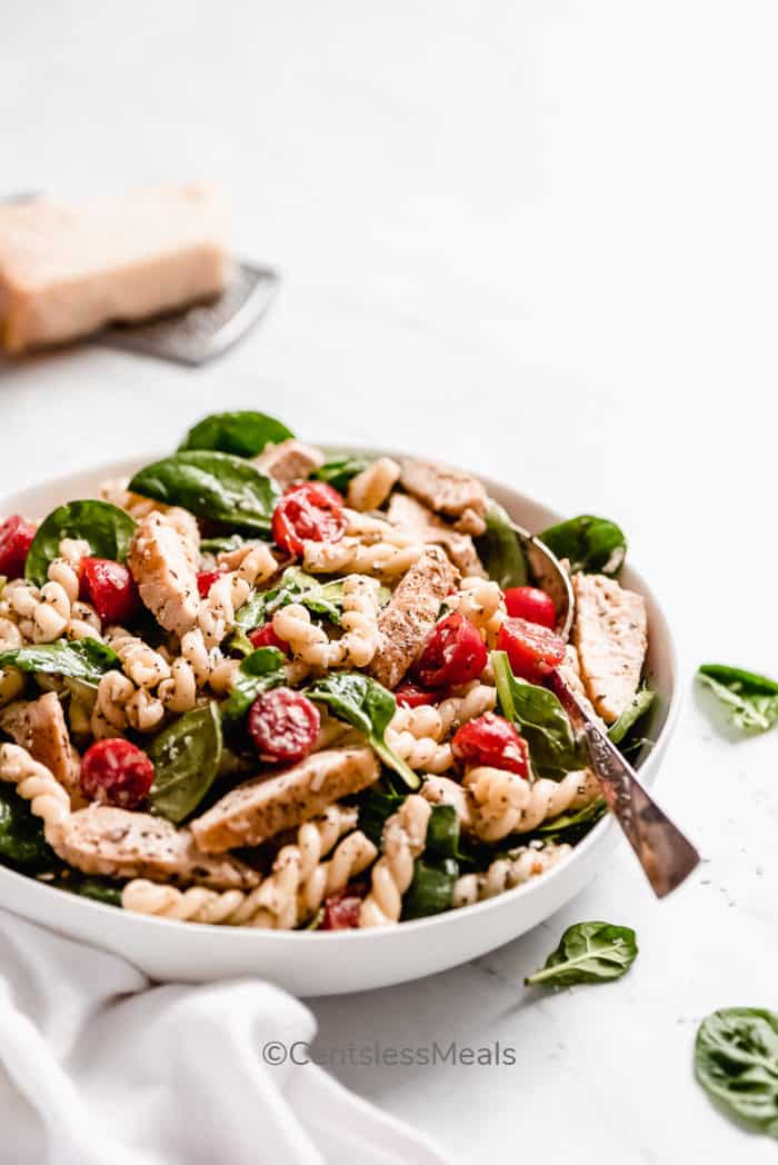 Spinach and Tomato Chicken Pasta Salad in a large white bowl, with spinach leaves on the side.