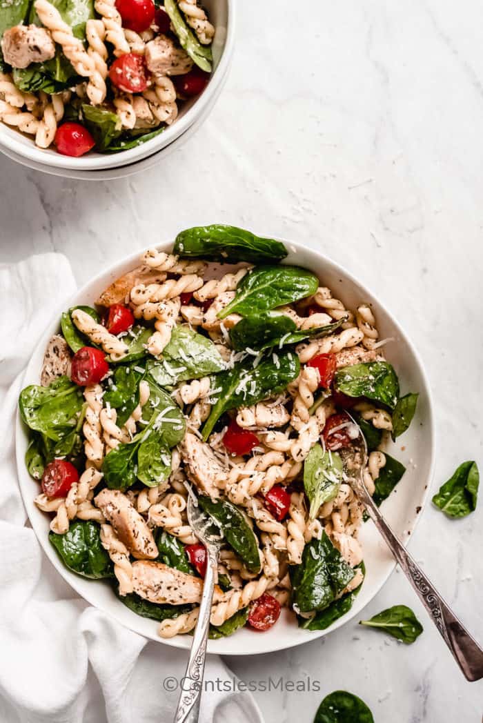 Spinach and Tomato Chicken Pasta Salad served in a white bowl with two silver spoons, garnished which parmesan cheese.