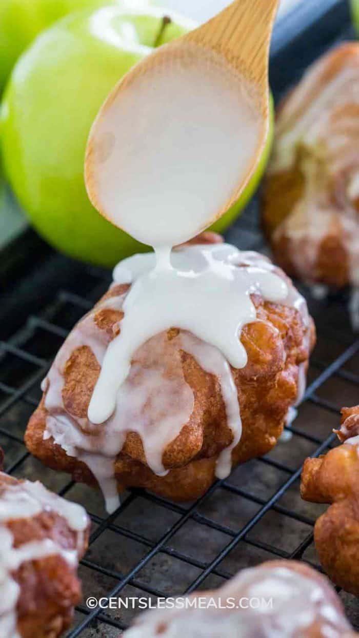 A white glaze being applied with a wooden spoon to Apple Fritters on a cooling rack
