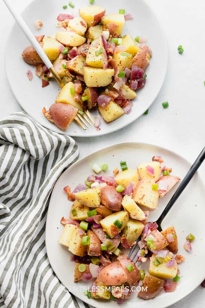 Warm German Potato Salad served on two white plates. One has a black fork while the other has a white and gold fork on a white background.