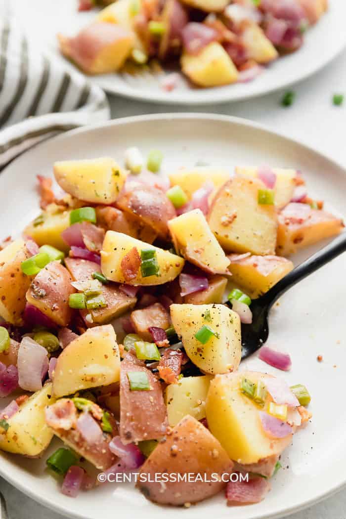 Warm German Potato Salad served on a white plate. A black fork is scooping up chunks of potato, bacon and red onion.