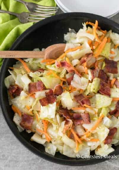 Fried cabbage and bacon in a black pan with a wooden spoon