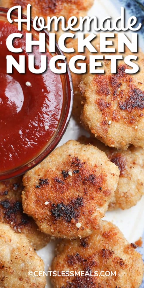 Homemade chicken nuggets served on a white plate with a bowl of ketchup, with writing.