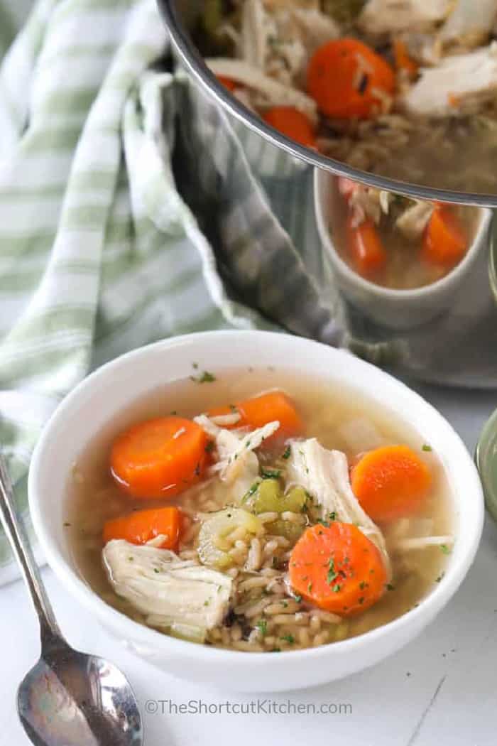 A pot of homemade chicken and rice soup with a white bowl of soup in front.