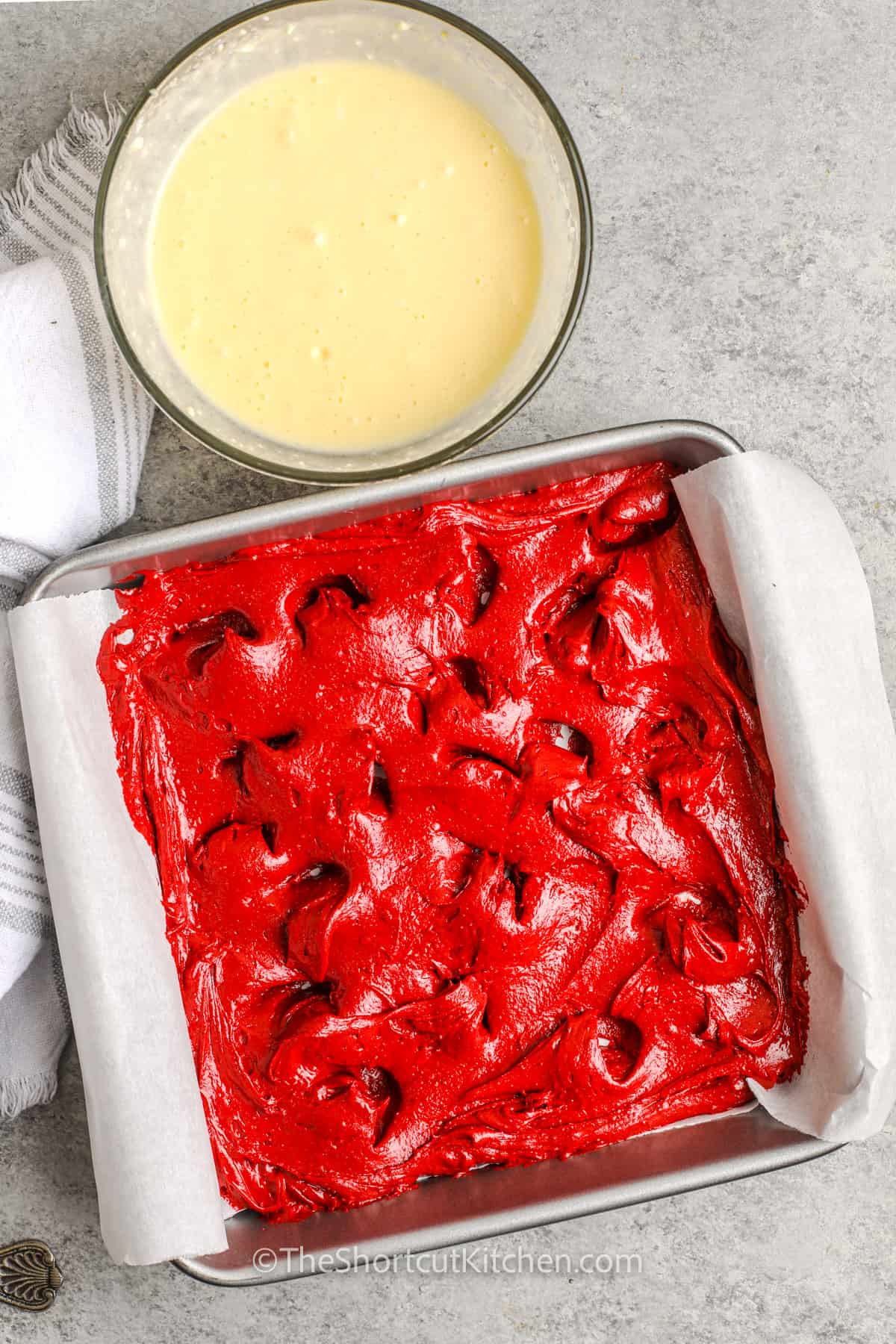 Red Velvet Brownie batter prepared in a pan with cheesecake mixture in a bowl beside it.