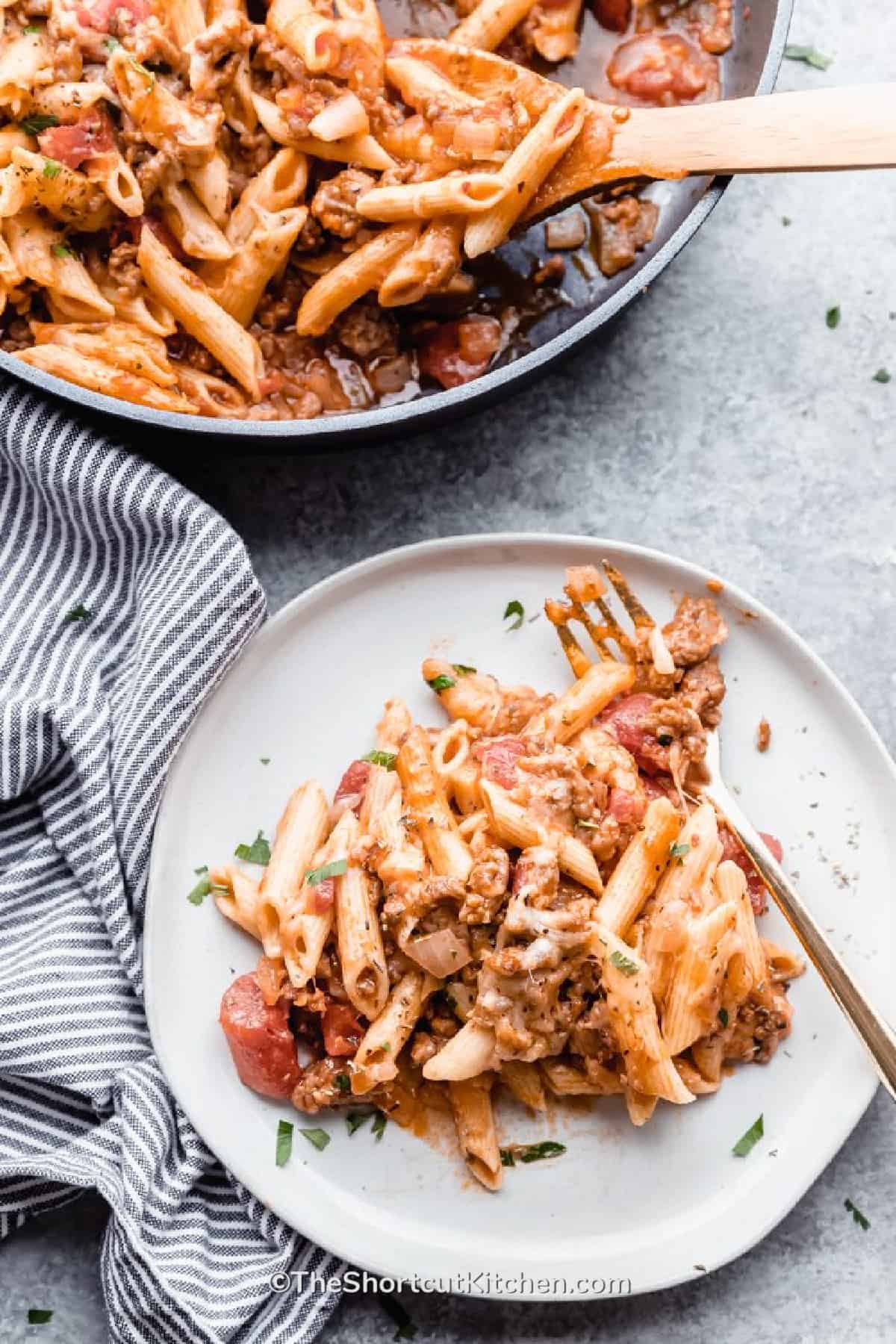 Italian Sausage Pasta and on a white plate with a fork, and the remaining pasta in a pan with a wooden spoon.