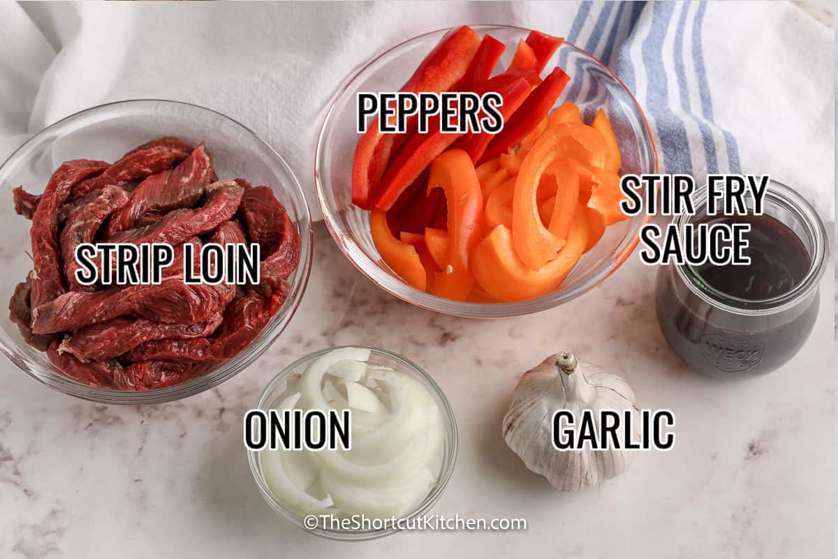 ingredients assembled to make steak stir fry, including steak, onion, peppers, stir fry sauce, garlic, and onion