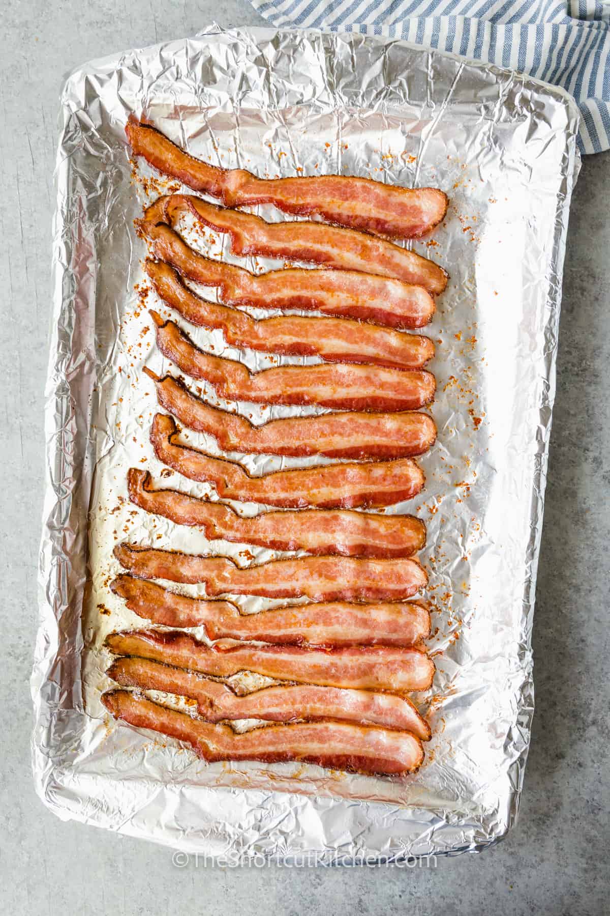 cooked oven baked bacon, lined up in a row, on a foiled lined baking sheet