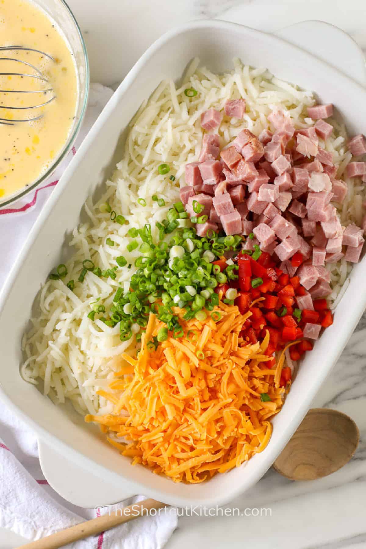 ham, hash brown potatoes, green onion, cheese and red peppers in a white casserole dish, with whisked eggs on the side, ready to make hash brown breakfast casserole