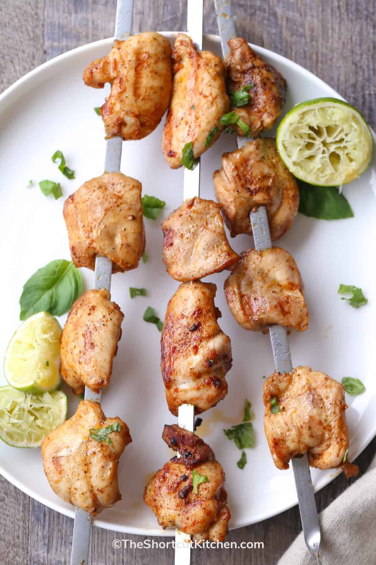 Skewered lime grilled chicken kabobs on a white plate with cilantro and lime wedges on the side.