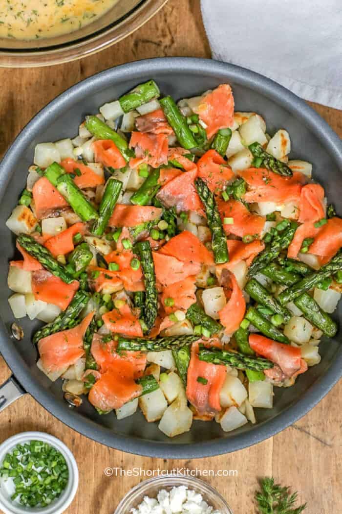 Salmon asparagus frittata in a frying pan with egg mixture, feta and chopped green onion on the side