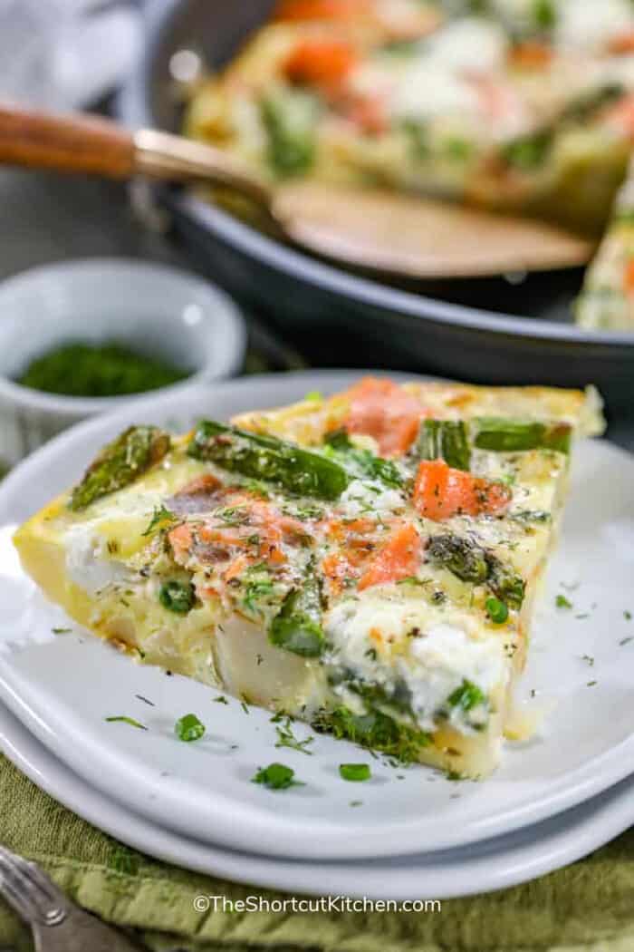 Salmon asparagus frittata on a small white plate, with a pan of salmon asparagus frittata in the background.