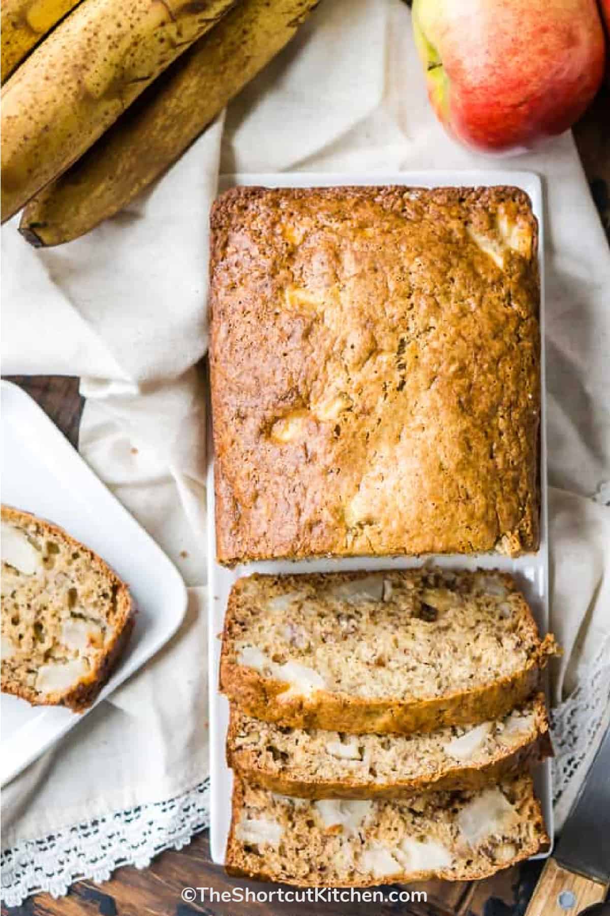 Apple banana bread on a white plate with some cut into slices with apples and bananas on the side