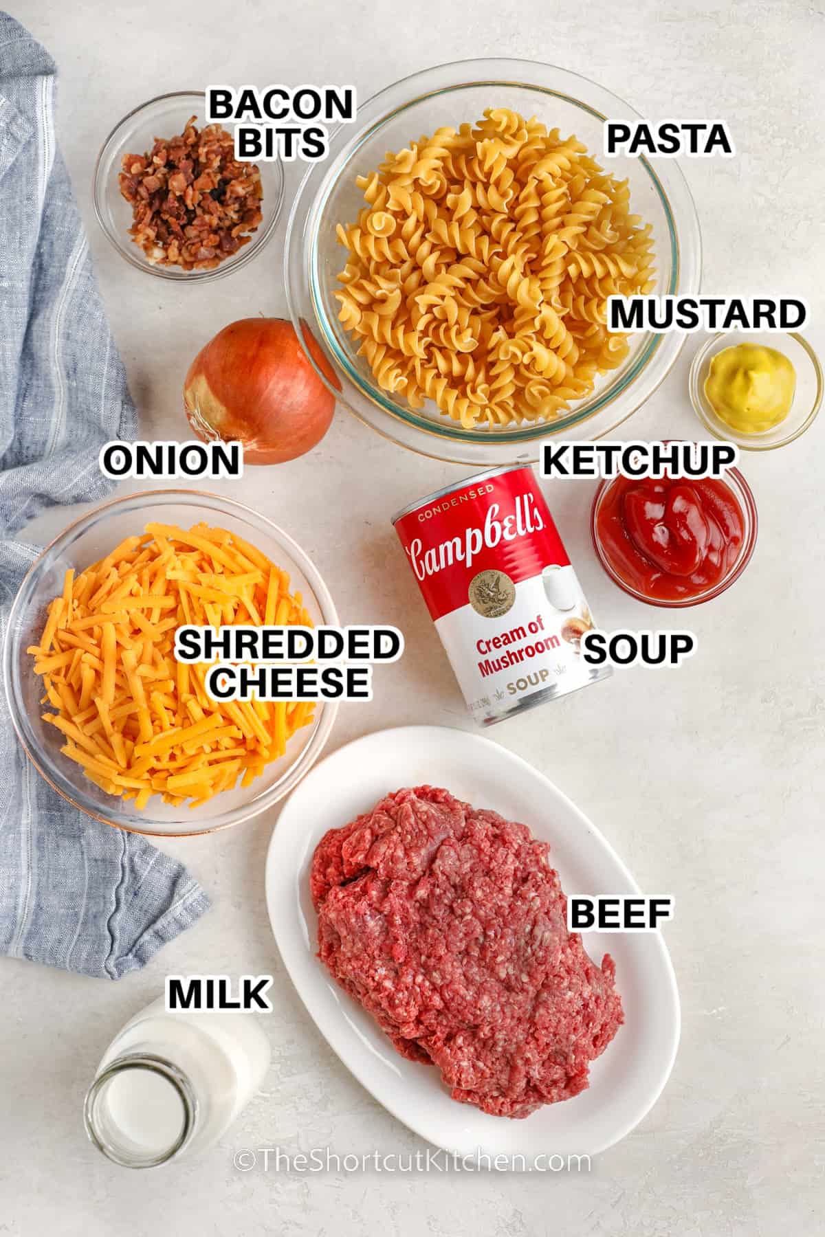 ingredients to make bacon cheeseburger casserole: bacon bits, pasta, mustard, ketchup, onion, shredded cheese, soup, beef, and milk