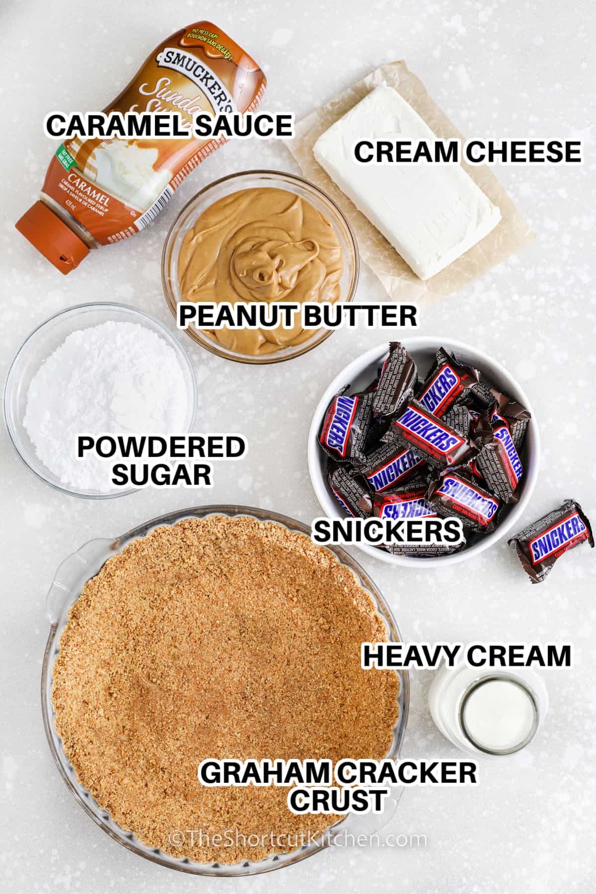 Ingredients to make Snickers Pie labeled: cream cheese, caramel sauce, peanut butter, powdered sugar, snickers, heavy cream, and a graham cracker crust