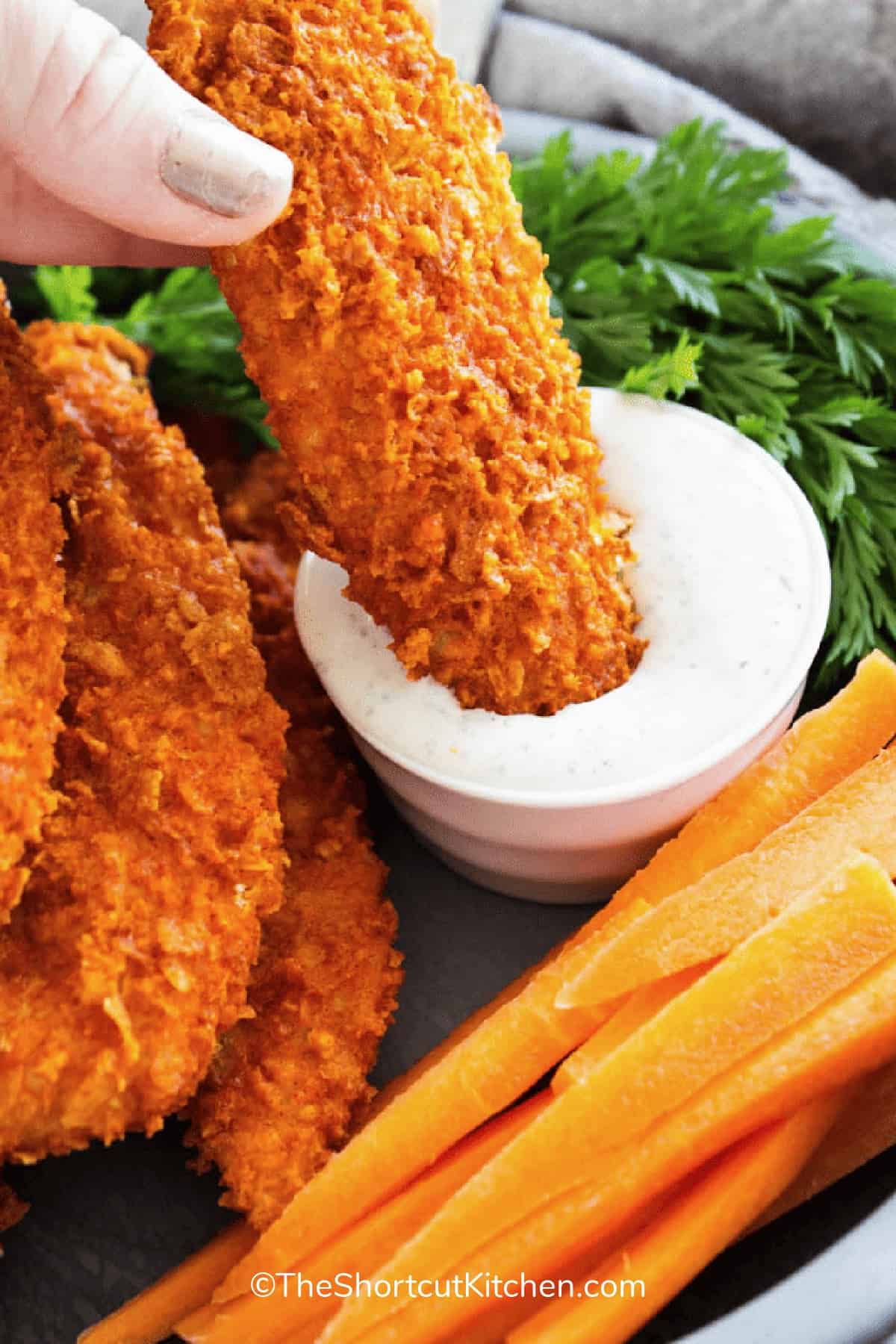 Crispy buffalo chicken tenders on a platter with carrot sticks, and one chicken tender is being dipped into dressing