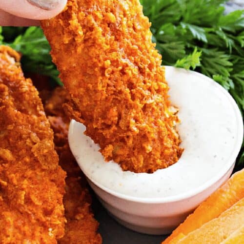 Close up of crispy buffalo chicken tenders on a platter with carrot sticks, and one chicken tender dipped into dressing