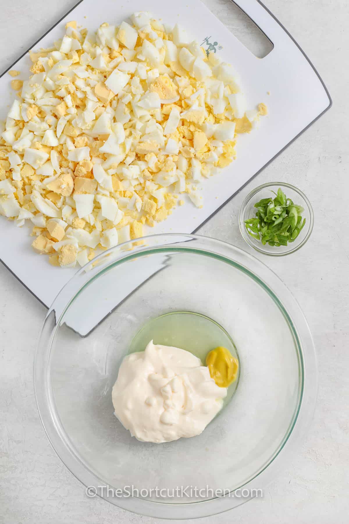 mayonnaise, mustard and pickle juice in a clear bowl, with chopped hard boiled eggs on a cutting board, and green onion in a tiny bowl, assembled to make the best egg salad recipe