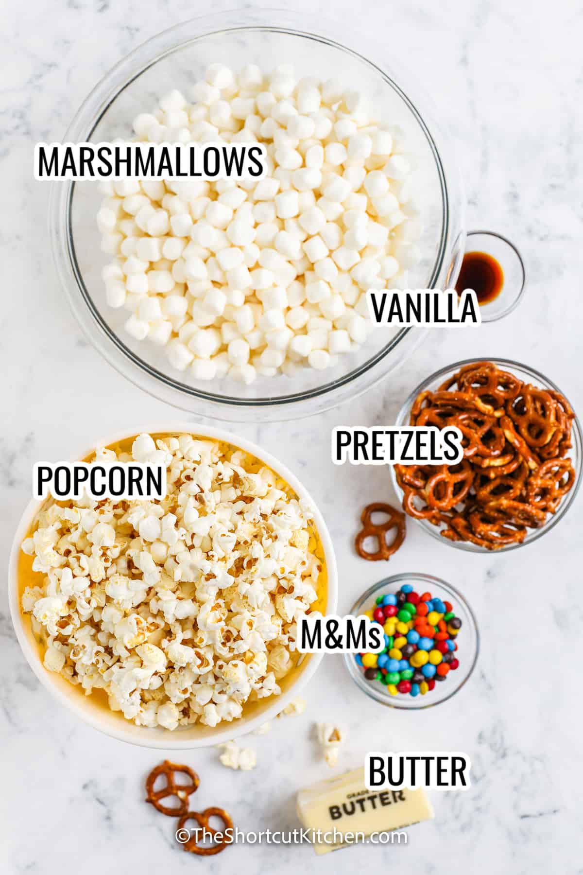 ingredients assembled to make popcorn bars, with labels including popcorn, marshmallows, pretzels, M&Ms, and butter