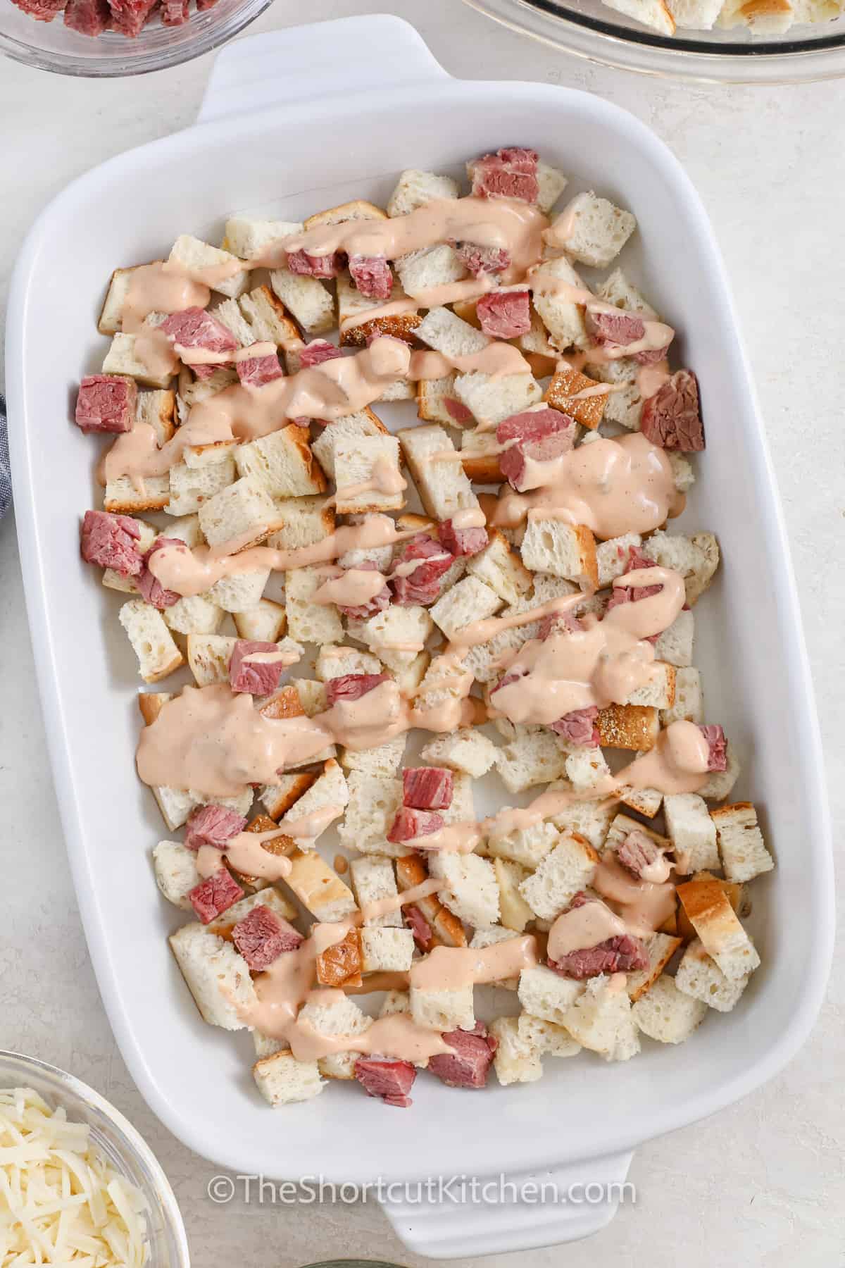 Looking for an easy comfort dinner? This Reuben casserole is layered with buttery rye bread, diced corned beef, drained sauerkraut, and gooey Swiss cheese, then baked in a 9×13 dish. Add Thousand Island and pickles on top for true deli flavor. A family-friendly easy Reuben casserole that doubles as a great make-ahead meal. #theshortcutkitchen adding thousand island to bread and meat to make Reuben Casserole