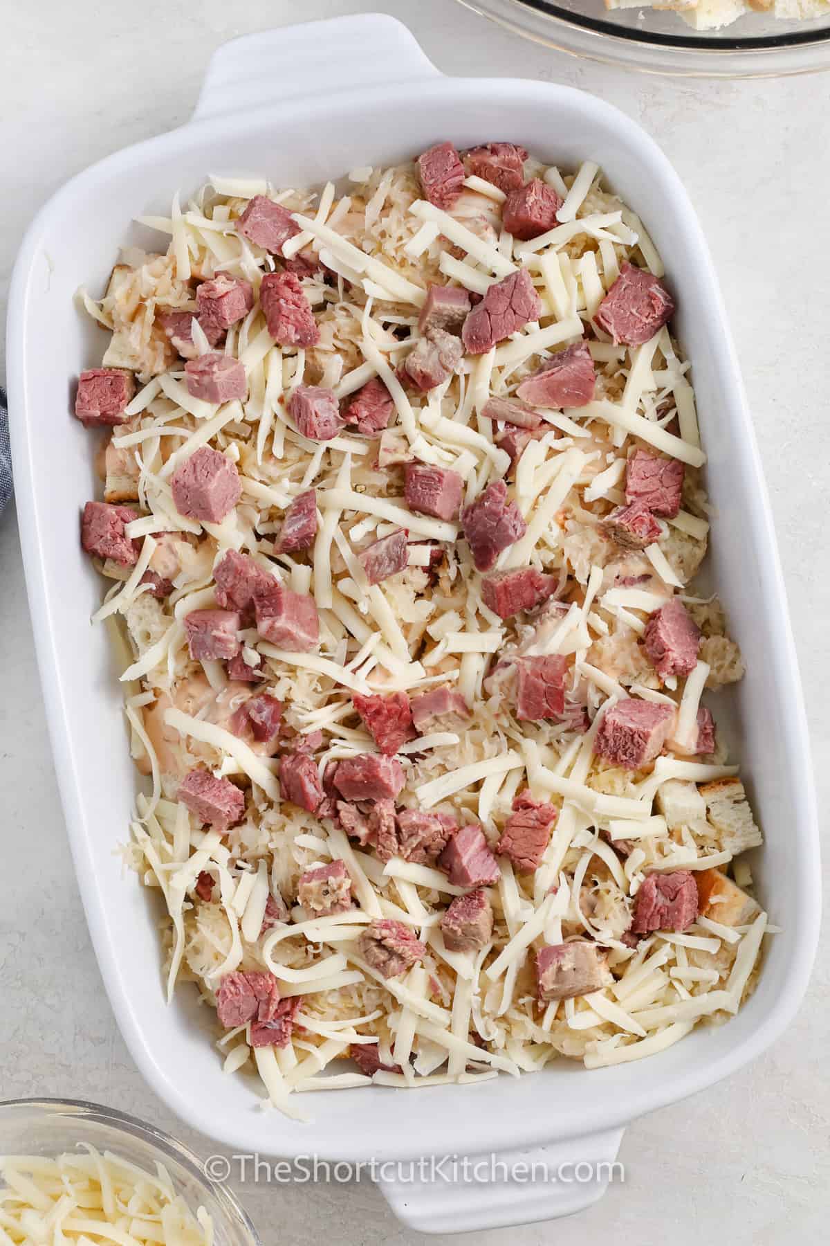 Skip the skillet and make a baked Reuben casserole instead. This easy Reuben casserole layers rye bread, corned beef, sauerkraut, and melty Swiss with creamy Thousand Island for a tangy, cheesy bite every time. It’s a simple corned beef casserole that’s perfect for potlucks, game day, or a weeknight comfort-food dinner. #theshortcutkitchen adding cheese to casserole dish to make Reuben Casserole