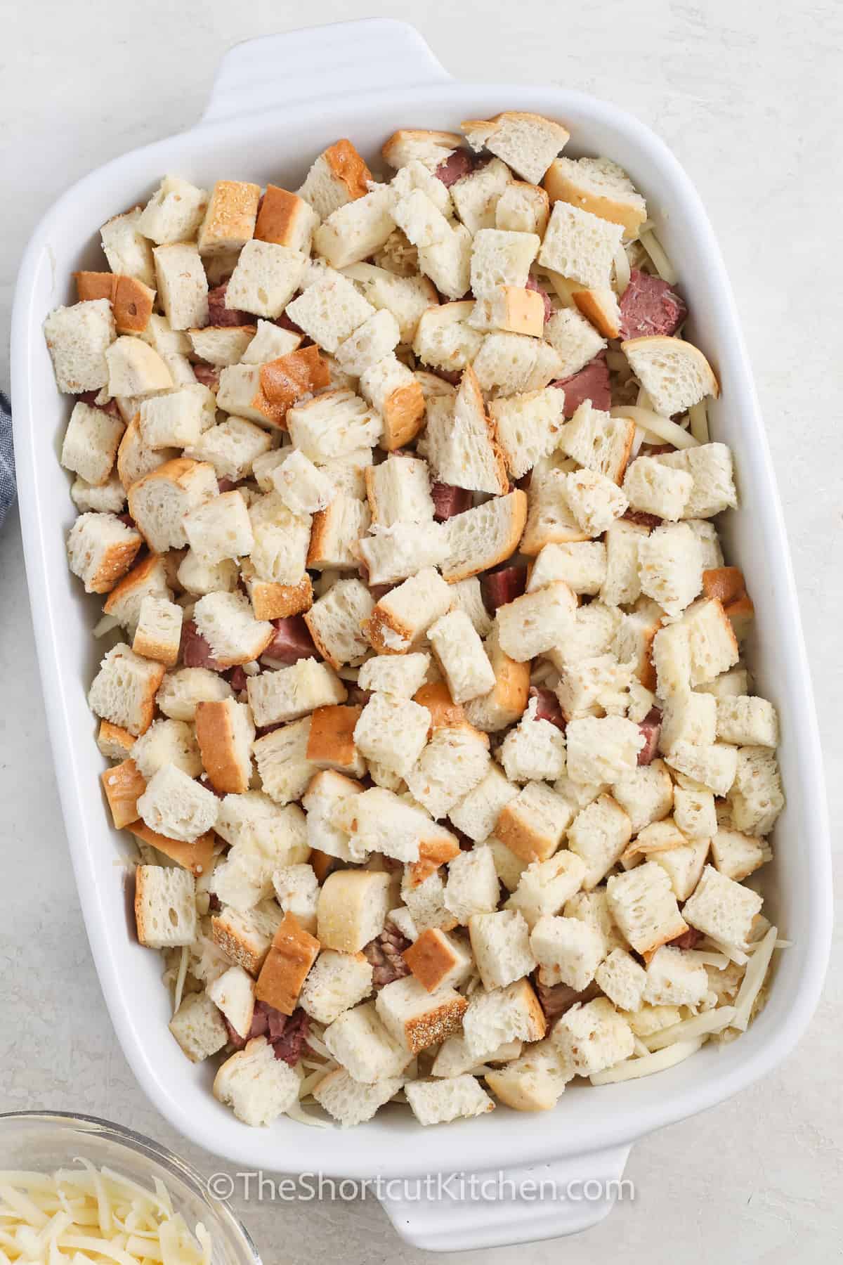 This Reuben casserole has all the classic deli flavors baked into one easy dinner. Buttery rye bread cubes are layered with diced corned beef, well-drained sauerkraut, and plenty of Swiss cheese, then finished with Thousand Island dressing and chopped pickles. A cozy Reuben casserole recipe that bakes up bubbly in a 9×13 pan and feeds a hungry crowd. #theshortkitchen adding bread on top of ingredients in the dish to make Reuben Casserole