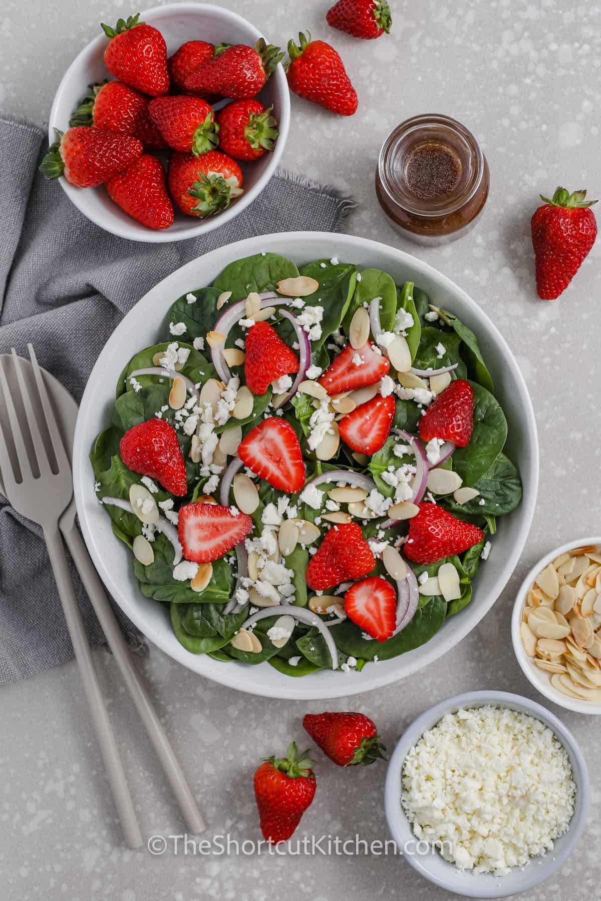 Spinach and Strawberry Salad in a white bowl surrounded by feta cheese, almonds, strawberries and dressing