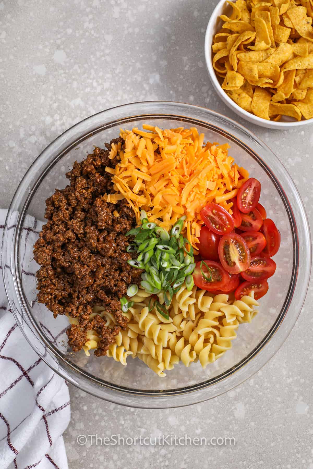 Taco pasta salad is a fun twist on classic taco salad with rotini pasta, seasoned beef, cheddar cheese, lettuce, tomatoes, and Catalina dressing. It is quick to prep, easy to customize, and perfect for summer gatherings or busy weeknight dinners. Add black beans, corn, olives, cilantro, or sour cream to make this taco pasta salad recipe your own. #theshortcutkitchen ingredients in a bowl to make Taco Pasta Salad