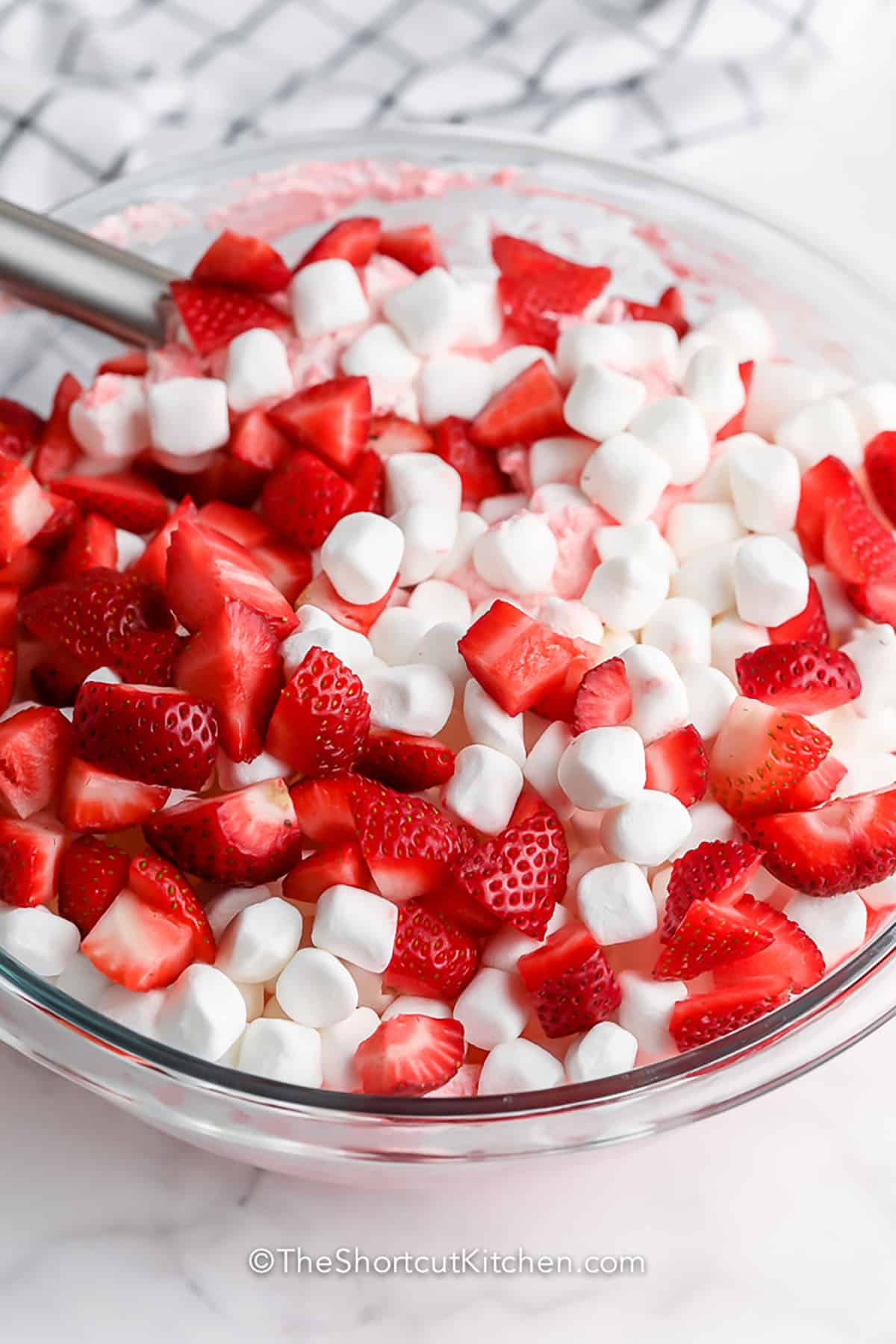 This easy strawberry fluff recipe turns a few simple ingredients into a cool and creamy dessert salad everyone loves. Made with strawberry Jello, yogurt, Cool Whip, mini marshmallows, and strawberries, it is a quick no-bake side dish or dessert for potlucks and parties. The optional crushed pineapple adds even more fruit flavor and a classic fluff salad twist. #theshortcutkitchen adding strawberries and marshmallows to ingredients to make Strawberry Fluff