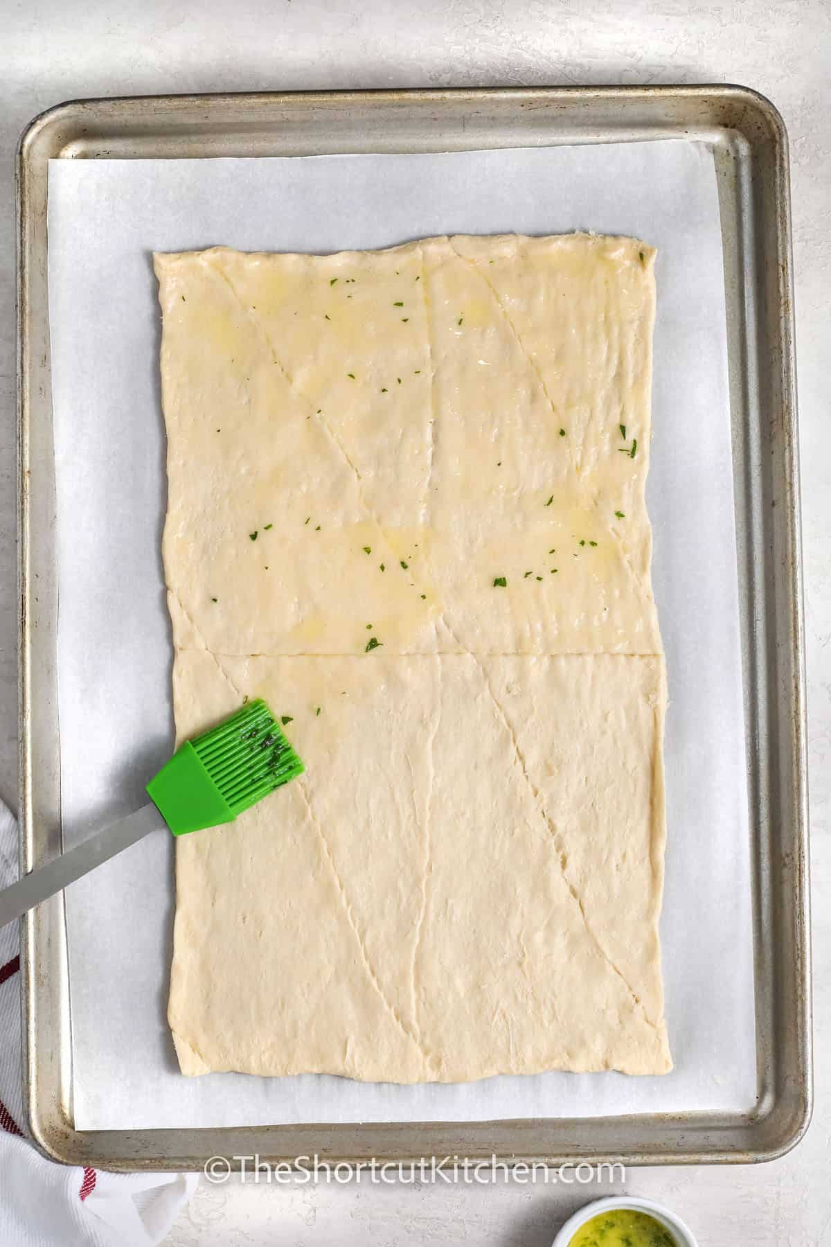 Crescent Roll Pizza makes dinner easy when time is short and everyone is hungry. Crescent dough bakes into a buttery, flaky crust with no kneading or rising, so you can get it in the oven fast. Top it with pizza sauce, mozzarella, and pepperoni, or use whatever toppings you have on hand. Everything bakes on one sheet pan, which means less prep, quick serving, and a faster cleanup. #theshortcutkitchen adding garlic butter to crescent roll dough to make Crescent Roll Pizza