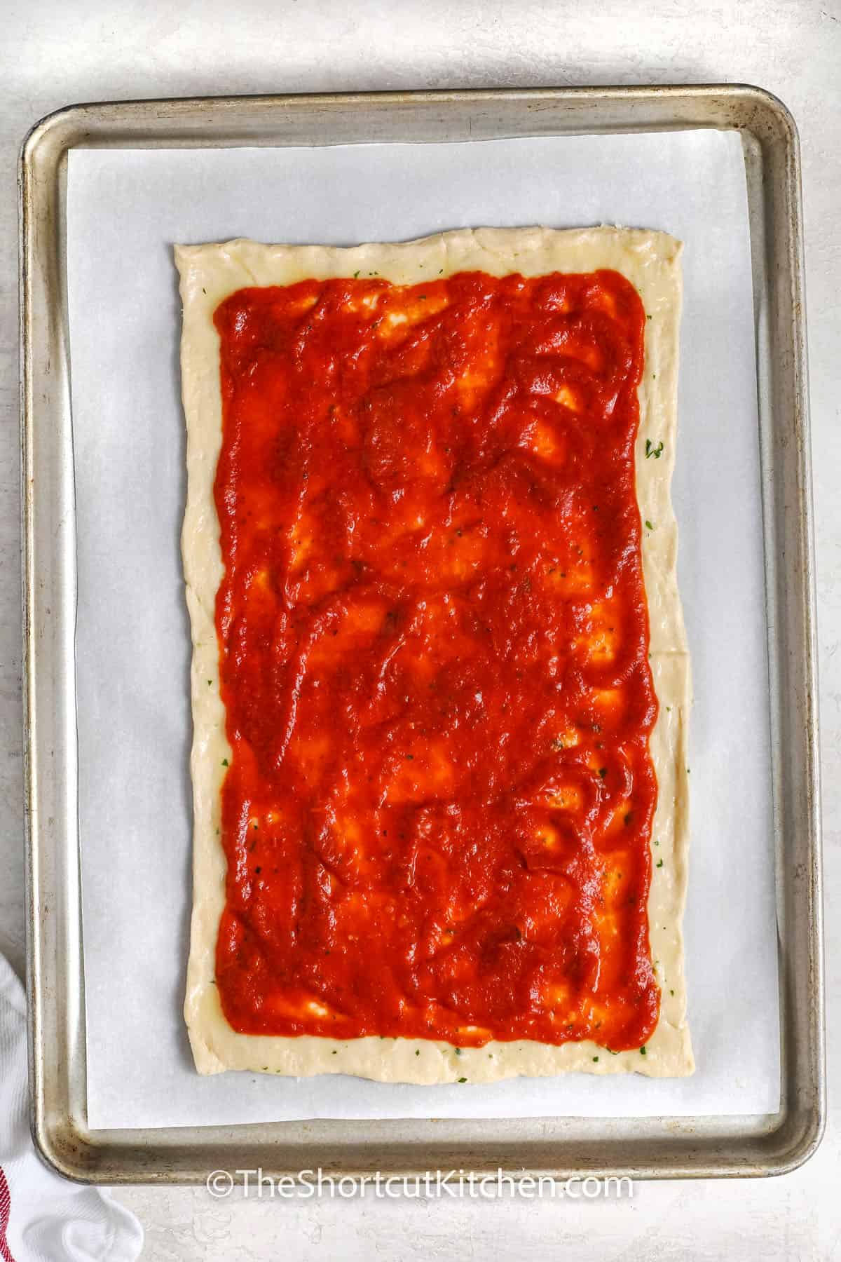This crescent roll pizza recipe features crispy, garlicky slices of pizza ready in under 30 minutes—it's the best for a quick meal or appetizer when you're in a hurry. Just roll out a can of crescent roll dough, then brush on garlic butter. Add pizza sauce, mozzarella cheese, pepperoni, and your favorite toppings. Bake until the crust is golden brown and serve hot. #theshortcutkitchen adding pizza sauce to crescent roll dough to make Crescent Roll Pizza