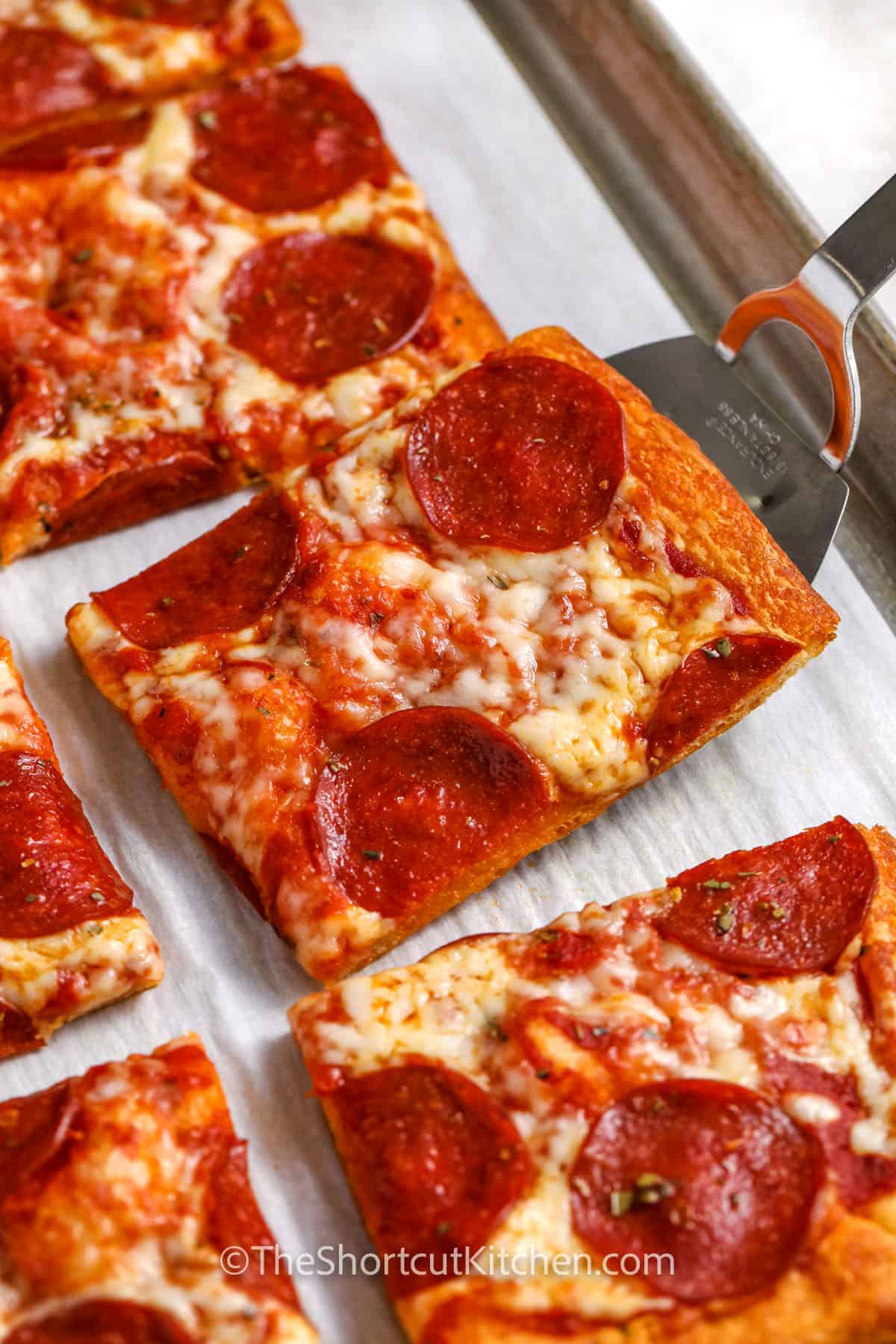 Crescent Roll Pizza is the ultimate shortcut recipe for pizza night. Unroll crescent roll dough, pinch the seams, brush with garlic butter, then add sauce, mozzarella, and pepperoni. This crescent roll pizza recipe cooks quickly on a sheet pan and slices into neat squares for serving. Customizable toppings make it great for families, parties, and last-minute meals. Try serving it with ranch dipping sauce for an extra special appetizer. #theshortcutkitchen taking a slice of Crescent Roll Pizza off the sheet pan